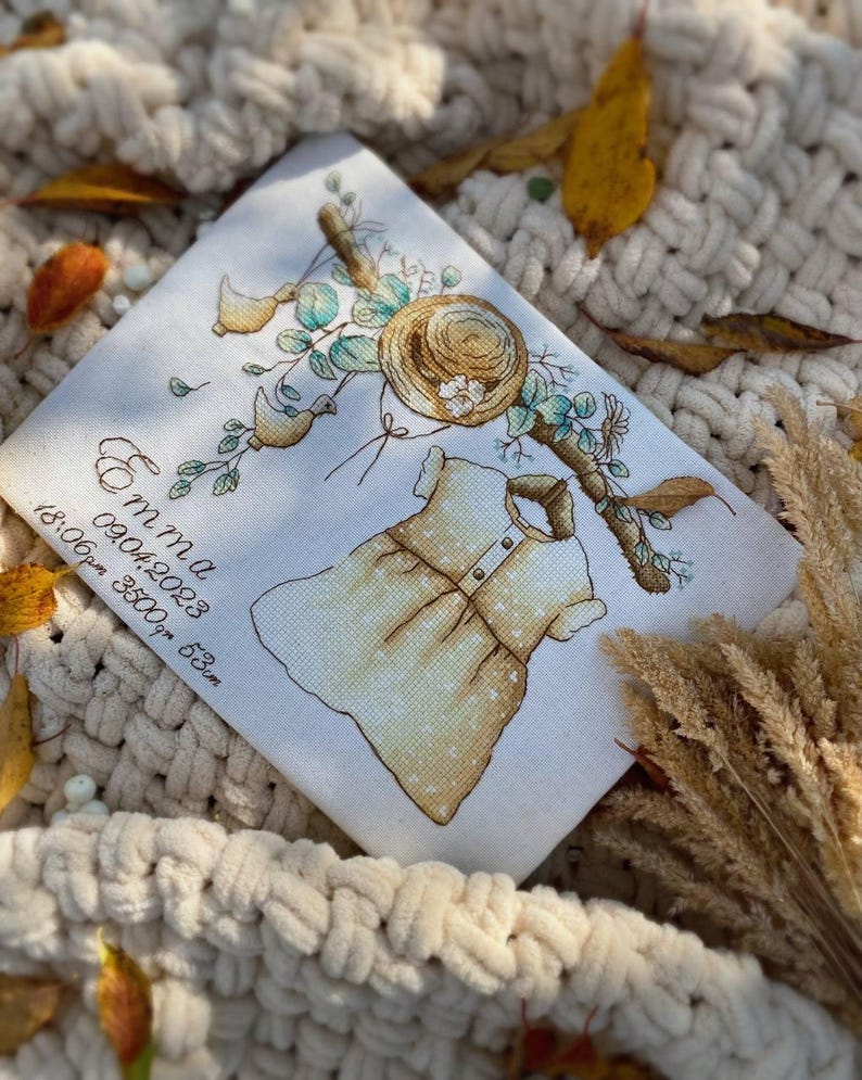 Peut inclure: Un panneau en tissu blanc avec une broderie au point de croix repr&eacute;sentant une robe de b&eacute;b&eacute; jaune, un chapeau de paille et des feuillages. Le texte indique Emma, 09.04.2023, 18:06pm, 3500g, 53cm. Broderie aux tons jaunes, verts et bruns.
