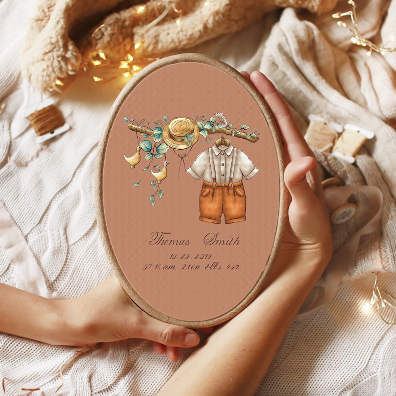 Op de afbeelding: Ovale borduurring met een aquarel illustratie van een babyoutfit, een strohoed en decoratieve elementen. De tekst luidt "Thomas Smith" en de geboortedatum en -tijd. Het kleurenpalet omvat tinten bruin, oranje en groen.
