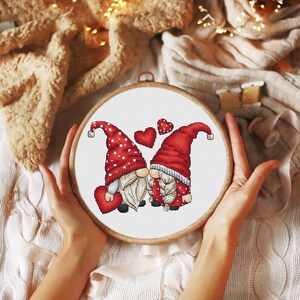 May include: Cross-stitch embroidery hoop featuring a design of two gnomes holding hands and hearts. The gnomes are wearing red hats with white polka dots and have white beards.