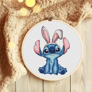May include: Cross stitch pattern of a blue Stitch character wearing pink bunny ears. The pattern is framed in a wooden embroidery hoop.