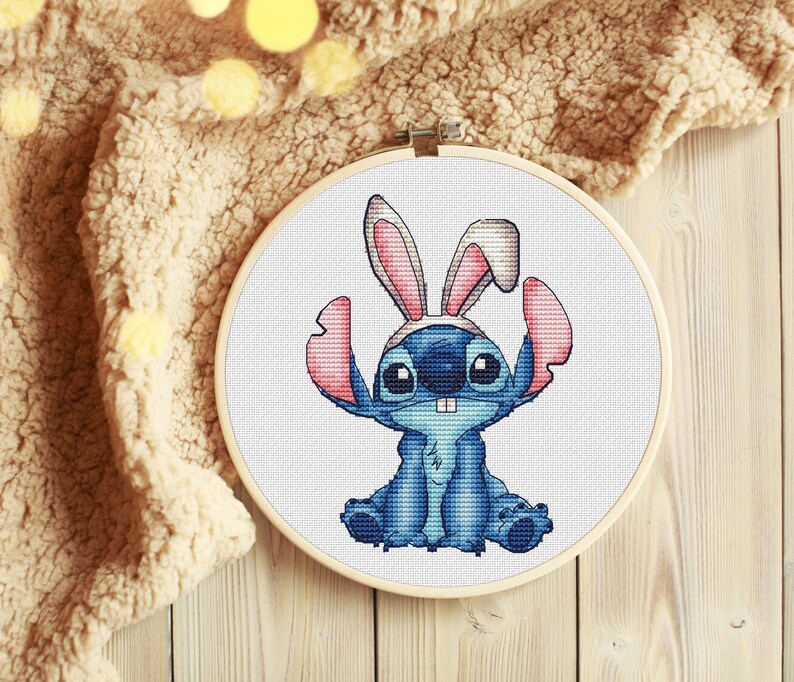 May include: Cross stitch pattern of a blue Stitch character wearing pink bunny ears. The pattern is framed in a wooden embroidery hoop.