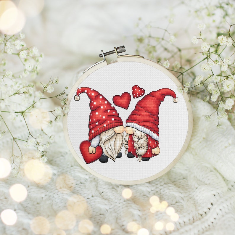 May include: Cross-stitch pattern of two gnomes kissing with red hearts around them. The gnomes are wearing red hats with white polka dots and white beards.