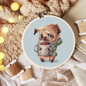 May include: A cross-stitch embroidery project in a circular wooden hoop featuring a tan pug dog wearing a green hooded sweatshirt and holding a large, polka-dotted mug filled with a dark beverage, with its tongue sticking out into the drink.