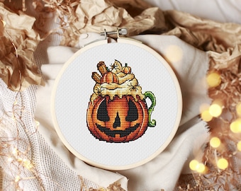 May include: A cross-stitch pattern of a pumpkin-shaped mug with whipped cream, cinnamon sticks, and pumpkin spice. The mug has a jack-o'-lantern face.