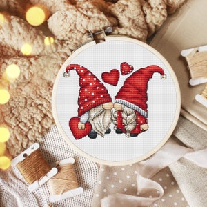 May include: Cross stitch pattern of two gnomes in red hats with white polka dots, kissing with hearts around them. The gnomes are holding red hearts.
