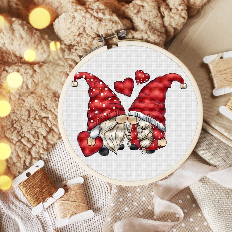 May include: Cross stitch pattern of two gnomes in red hats with white polka dots, kissing with hearts around them. The gnomes are holding red hearts.