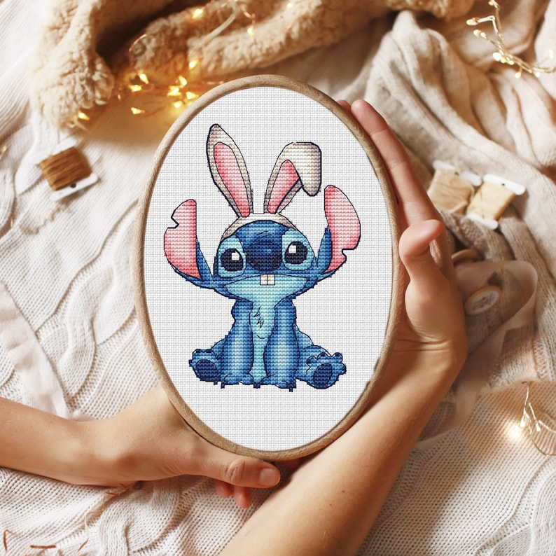 May include: Cross stitch embroidery hoop with a blue Stitch character wearing a pink bunny ear headband. The background is white.