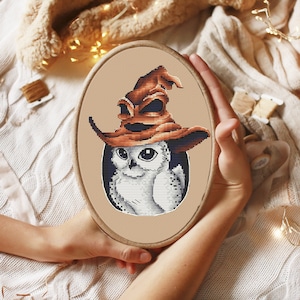 Op de afbeelding: Een ovale borduurring met een kruissteekpatroon van een witte uil die een grote, verweerde bruine tovenaarshoed draagt. De uil heeft grote zwarte ogen en gedetailleerd verenkleed op een effen beige stoffen achtergrond in het houten frame.
