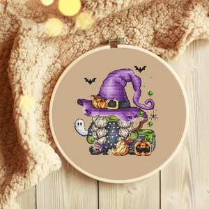 Pu&ograve; includere: Uno schema di punto croce di un gnomo strega viola e verde con un cappello nero, che tiene una scopa e una zucca. Lo gnomo &egrave; circondato da pipistrelli, ragni e un calderone.