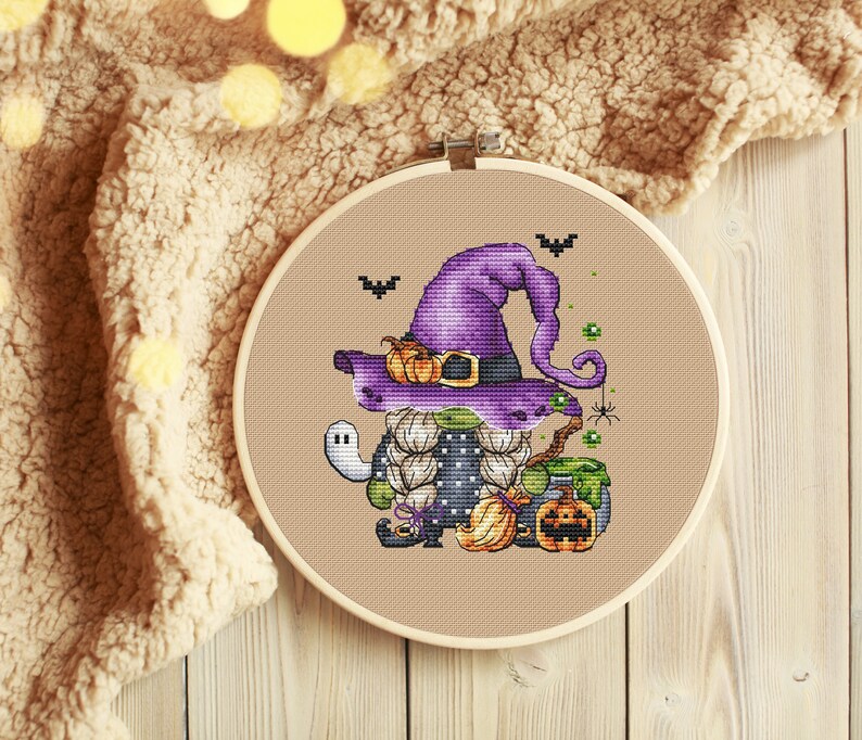 Pu&ograve; includere: Uno schema di punto croce di un gnomo strega viola e verde con un cappello nero, che tiene una scopa e una zucca. Lo gnomo &egrave; circondato da pipistrelli, ragni e un calderone.