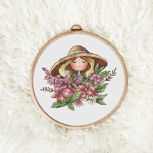 May include: A cross-stitch embroidery hoop with a white background featuring a cartoon girl with blonde hair wearing a brown and blue hat. The girl is surrounded by pink, white, and green flowers.