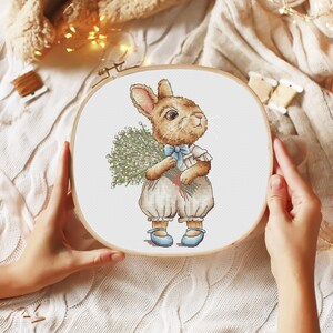 以下が含まれることがあります： 白い背景に、白い花束を持った茶色のウサギが刺繍されたクロスステッチの刺繍枠。ウサギは青いリボンと白いドレスを着ています。