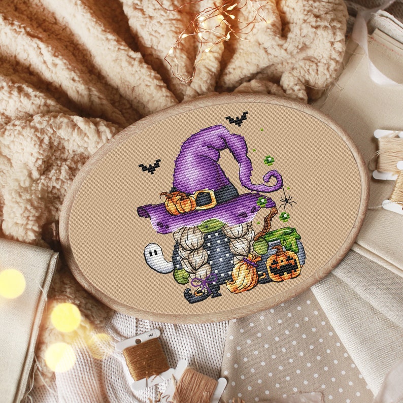 Pu&ograve; includere: Un ricamo a punto croce di un gnomo strega viola e verde che tiene un calderone e una zucca. Lo gnomo indossa un cappello viola con una fibbia nera e un vestito bianco e nero. Lo sfondo &egrave; un tessuto beige.