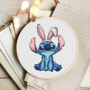 May include: A blue Stitch character from the Disney movie Lilo & Stitch wearing pink bunny ears. The character is sitting with its legs crossed and has a toothy grin. The image is a cross-stitch pattern.