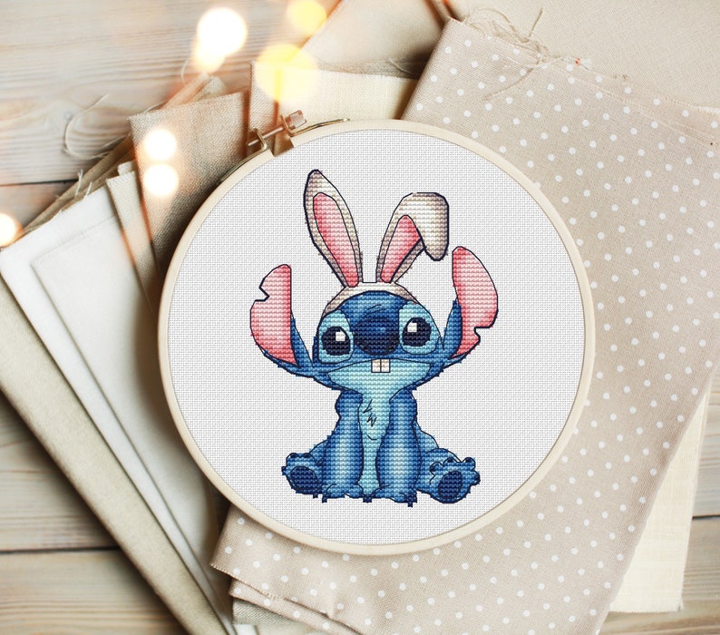 May include: A blue Stitch character from the Disney movie Lilo & Stitch wearing pink bunny ears. The character is sitting with its legs crossed and has a toothy grin. The image is a cross-stitch pattern.