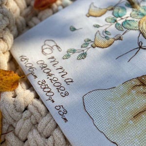 Peut inclure: Une pi&egrave;ce de tissu blanc avec une broderie au point de croix d&eacute;taill&eacute;e repr&eacute;sentant un arbre avec des oiseaux jaunes et des feuilles vertes. Le texte indique Emma, 09.04.2023, 18:06pm, 3500gm, 53cm. Inclut une forme jaune et des lettres brunes.