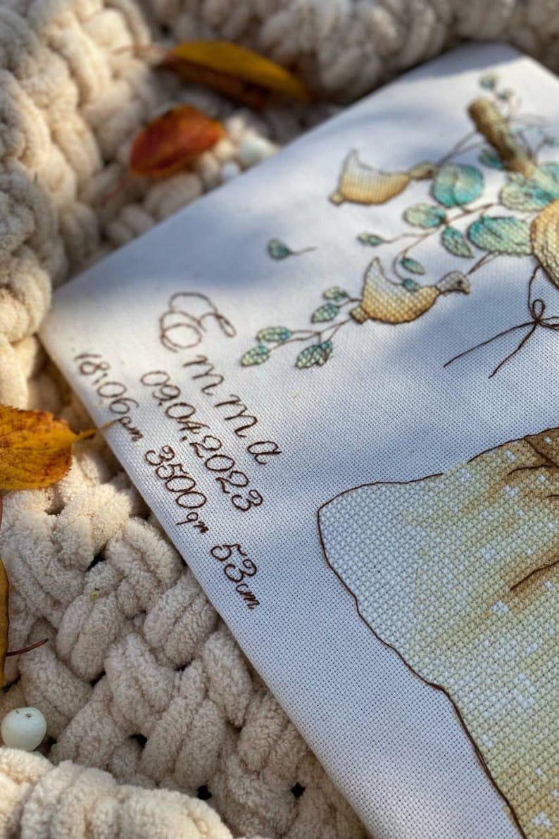 Peut inclure: Une pi&egrave;ce de tissu blanc avec une broderie au point de croix d&eacute;taill&eacute;e repr&eacute;sentant un arbre avec des oiseaux jaunes et des feuilles vertes. Le texte indique Emma, 09.04.2023, 18:06pm, 3500gm, 53cm. Inclut une forme jaune et des lettres brunes.