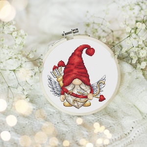 May include: A red and white gnome with angel wings holding a bow and arrow with hearts. The gnome is wearing a red hat with a white pom-pom. The gnome is on a white background.