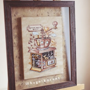 Op de afbeelding: Een ingelijst borduurwerk met kruissteek van een vintage koffiemolen met de tekst "Время пить кофе!" (Tijd om koffie te drinken!) in het Russisch. Het borduurwerk is op een beige achtergrond met bruine en gouden accenten. De lijst is van bruin hout met een verweerde afwerking. Het borduurwerk wordt getoond op een houten ezel.