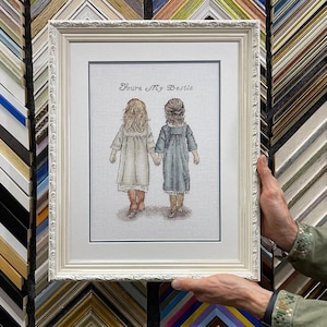 Op de afbeelding: Een ingelijst kunstwerk met twee meisjes die elkaars hand vasthouden, met de tekst "You're My Bestie" erboven. Het kunstwerk zit in een witte, sierlijke lijst. De meisjes dragen jurken en laarzen. De achtergrond is neutraal.