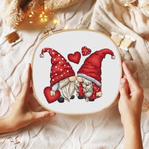 May include: Cross stitch pattern featuring two gnomes in red hats and white beards, holding red hearts and kissing. The gnomes are surrounded by red hearts.