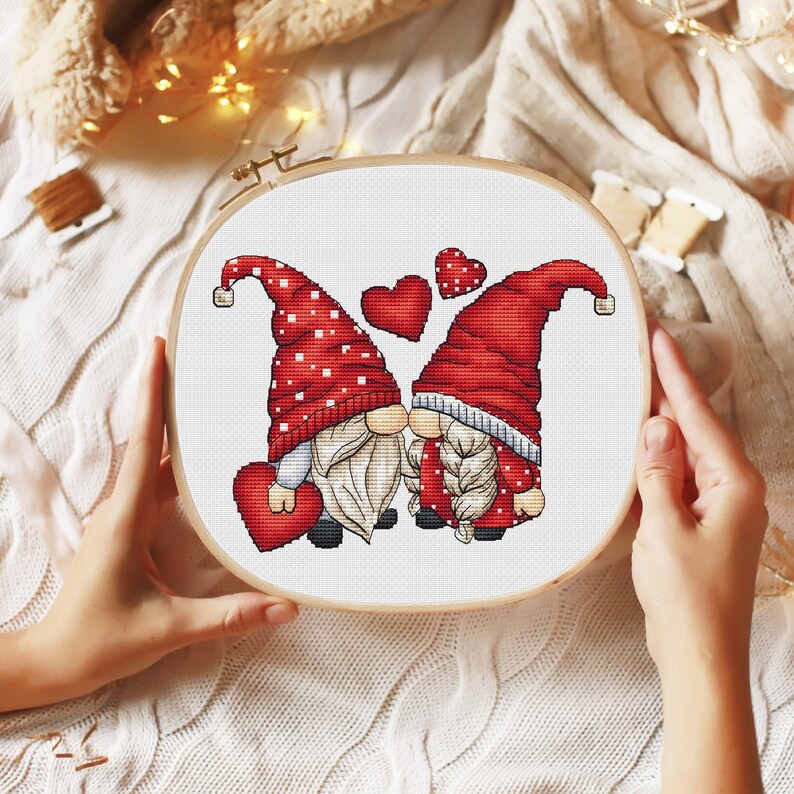 May include: Cross stitch pattern featuring two gnomes in red hats and white beards, holding red hearts and kissing. The gnomes are surrounded by red hearts.