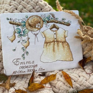 Peut inclure: Un panneau en tissu blanc avec une broderie au point de croix repr&eacute;sentant un chapeau de paille, une robe sur un cintre et deux oiseaux sur une branche. Le texte indique Emma, 09.04.2023, 18:06am, 3500gm, 53cm. Broderie aux fils marron, vert et jaune.