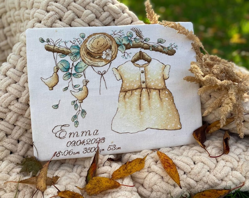 Peut inclure: Un panneau en tissu blanc avec une broderie au point de croix repr&eacute;sentant un chapeau de paille, une robe sur un cintre et deux oiseaux sur une branche. Le texte indique Emma, 09.04.2023, 18:06am, 3500gm, 53cm. Broderie aux fils marron, vert et jaune.