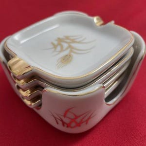 May include: A set of four square white ceramic ashtrays with gold trim and a gold wheat design. The ashtrays are stacked in a white holder with red accents. The set is designed for use as a decorative item.