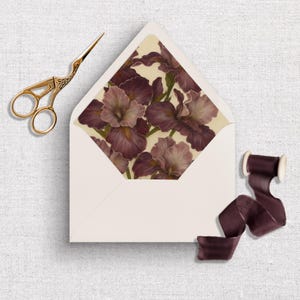 Op de afbeelding: Ivoorkleurige envelop met een bloemenirisontwerp aan de binnenkant. Gouden schaar en een spoel met donkerpaars lint liggen naast de envelop. De irissen zijn paars en lavendel.
