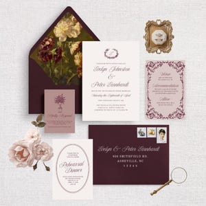 May include: A collection of wedding stationery in shades of burgundy, cream, and gold. Includes invitations, RSVP cards, and an envelope with a floral lining. A small framed ring and a magnifying glass are also present.