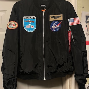 May include: Black bomber jacket with multiple NASA and space-themed patches, including a Space Lab J patch, a NASA patch, and a US flag patch. The jacket has a zipper closure and a red tag that says "Remove Before Flight".