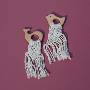 Può includere: Due morsi di legno con frange in macramé. Un morso ha la forma di un uccello e l'altro di una balena. Entrambi i morsi sono realizzati in legno naturale e hanno una frangia in macramé bianca.