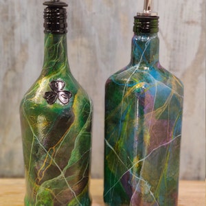 Pu&ograve; includere: Due bottiglie di vetro verde con disegni di vernice decorativi e beccucci di versamento neri e argentati. La bottiglia a sinistra ha un design a trifoglio.