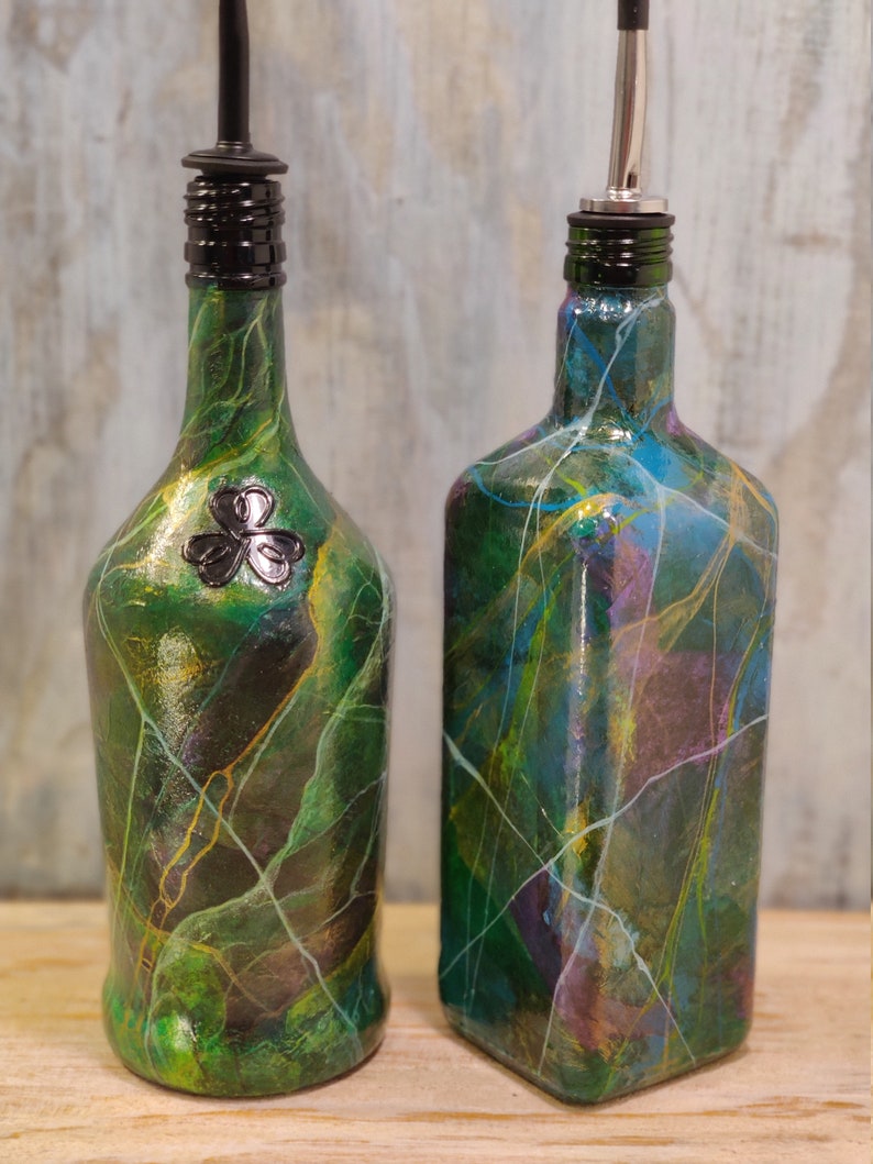 Pu&ograve; includere: Due bottiglie di vetro verde con disegni di vernice decorativi e beccucci di versamento neri e argentati. La bottiglia a sinistra ha un design a trifoglio.