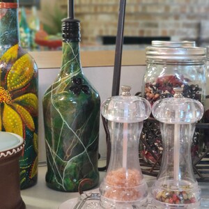 Pu&ograve; includere: Una collezione di bottiglie di vetro con vernice decorativa, tra cui una bottiglia verde con un design a trifoglio, una bottiglia gialla con un design floreale e due saliere e pepiere in vetro trasparente. Le bottiglie sono su un bancone con una superficie in legno marrone. Ci sono anche tre barattoli di vetro con coperchi, pieni di erbe e spezie essiccate.