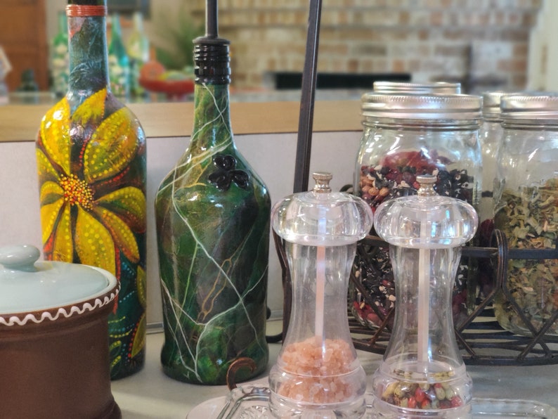 Pu&ograve; includere: Una collezione di bottiglie di vetro con vernice decorativa, tra cui una bottiglia verde con un design a trifoglio, una bottiglia gialla con un design floreale e due saliere e pepiere in vetro trasparente. Le bottiglie sono su un bancone con una superficie in legno marrone. Ci sono anche tre barattoli di vetro con coperchi, pieni di erbe e spezie essiccate.