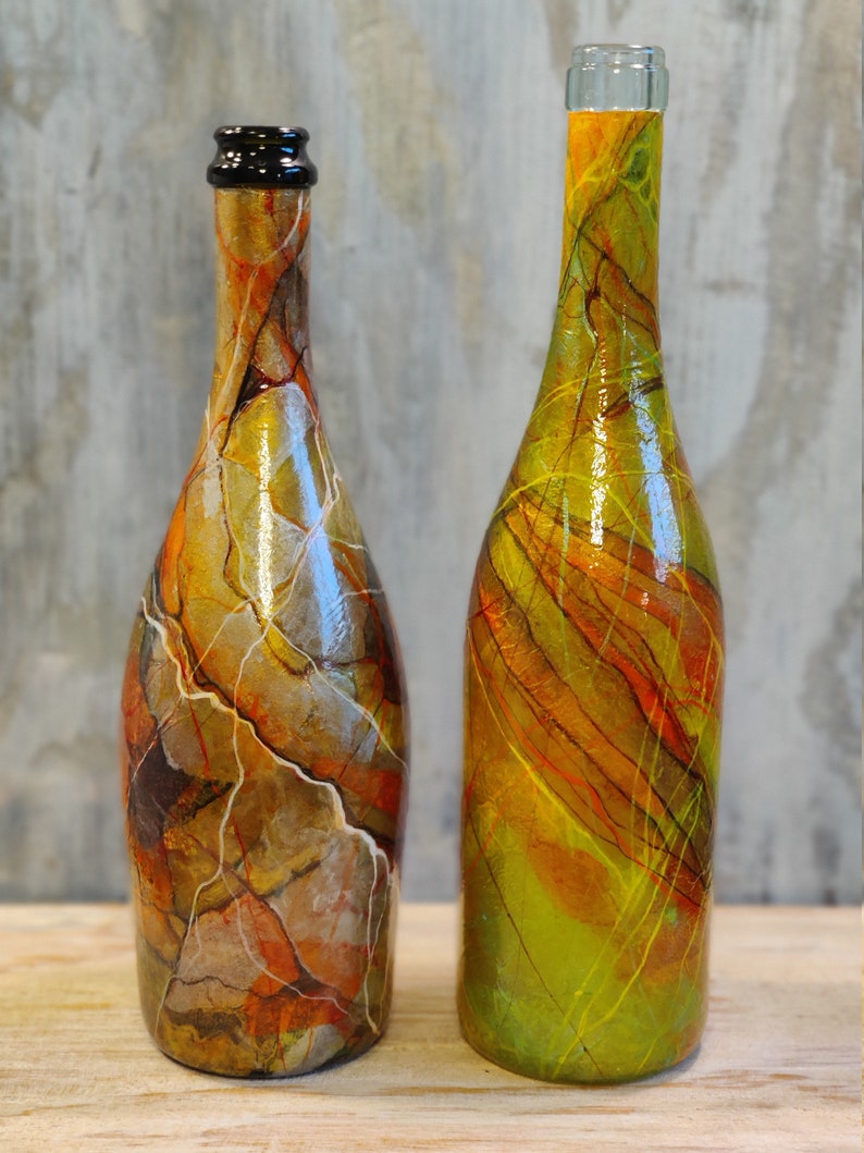 Pu&ograve; includere: Due bottiglie di vetro con disegni astratti colorati. Una bottiglia ha un design marmoreo marrone, arancione e bianco. L'altra bottiglia ha un design verde, giallo e arancione con linee sottili.