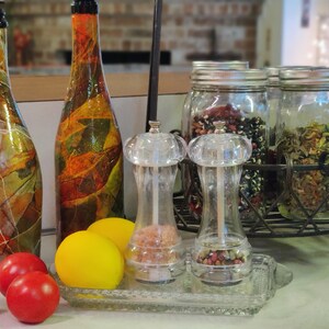Pu&ograve; includere: Due bottiglie di vetro decorative con disegni colorati, due macinini per sale e pepe in vetro trasparente e tre barattoli di vetro con coperchi pieni di vari ingredienti. I barattoli sono su una rastrelliera in metallo.