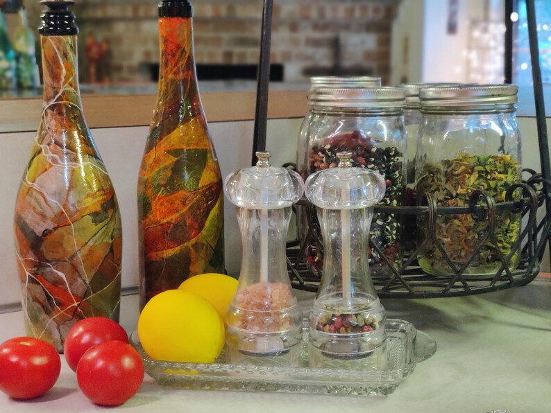 Pu&ograve; includere: Due bottiglie di vetro decorative con disegni colorati, due macinini per sale e pepe in vetro trasparente e tre barattoli di vetro con coperchi pieni di vari ingredienti. I barattoli sono su una rastrelliera in metallo.