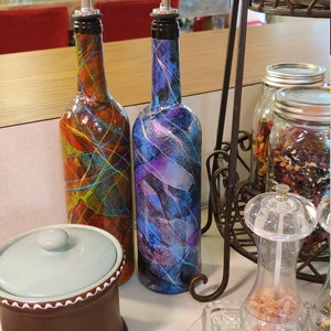 Pu&ograve; includere: Due bottiglie di vetro con disegni astratti colorati e beccucci versatori argentati. Le bottiglie sono su una superficie di legno con un contenitore in ceramica e un saliera di vetro.