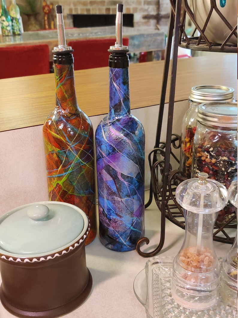 Pu&ograve; includere: Due bottiglie di vetro con disegni astratti colorati e beccucci versatori argentati. Le bottiglie sono su una superficie di legno con un contenitore in ceramica e un saliera di vetro.