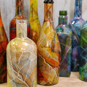 Pu&ograve; includere: Una collezione di otto bottiglie di vetro, ciascuna dipinta con un design marmorizzato unico in vari colori, tra cui arancione, giallo, blu, verde e viola. Le bottiglie sono disposte in fila su una superficie di legno.
