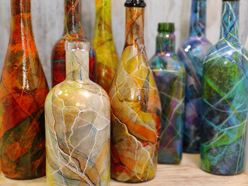 Pu&ograve; includere: Una collezione di otto bottiglie di vetro, ciascuna dipinta con un design marmorizzato unico in vari colori, tra cui arancione, giallo, blu, verde e viola. Le bottiglie sono disposte in fila su una superficie di legno.