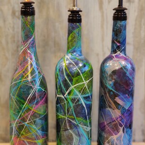 Pu&ograve; includere: Tre bottiglie di vetro con disegni astratti colorati e beccucci neri. Le bottiglie sono dipinte in tonalit&agrave; di blu, verde, viola e bianco.