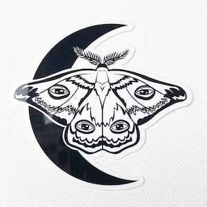 May include: Black and white sticker of a moth with multiple eyes, perched on a crescent moon.