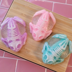 May include: Three frog-shaped soaps in shades of purple, pink, and teal, each with a translucent body and a white skeleton design. The soaps are arranged on a light brown wooden board.