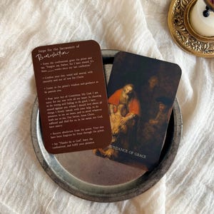 May include: A still-life image featuring two cards with religious text and artwork, resting on a small, round, silver tray. One card has text about the Sacrament of Reconciliation. The other card displays a painting with the words "ABUNDANCE OF GRACE". A decorative gold-colored candle holder is in the background.