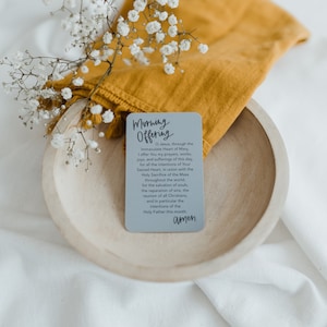 May include: A gray card with the text "Morning Offering" printed in black. The card is sitting on a wooden bowl with a yellow cloth draped over the side. The text on the card reads: "O Jesus, through the Immaculate Heart of Mary, I offer You my prayers, works, joys, and sufferings of this day, for all the intentions of Your Sacred Heart, in union with the Holy Sacrifice of the Mass throughout the world, for the salvation of souls, the reparation of sins, the reunion of all Christians, and in particular the intentions of the Holy Father this month. Amen."