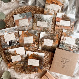 May include: A collection of affirmation cards featuring cottage scenes with text overlays, arranged on a woven mat. The cards have a vintage aesthetic, with soft colors and detailed illustrations. A brown envelope with the text "cottage of my heart" is also included.